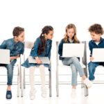four kids in denim clothes sitting on chairs and using laptops on white